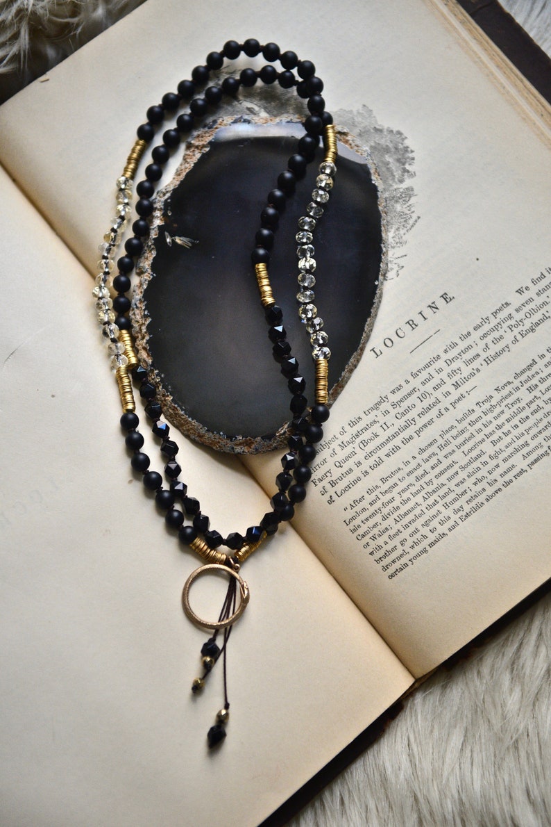 May include: A black and gold beaded necklace with a circular pendant. The necklace is lying on an open book with the text "LOCRINE" visible.