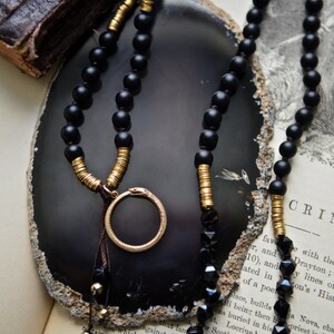May include: A black and gold beaded necklace with a circular pendant. The necklace is displayed on a grey agate slice and a book with text.