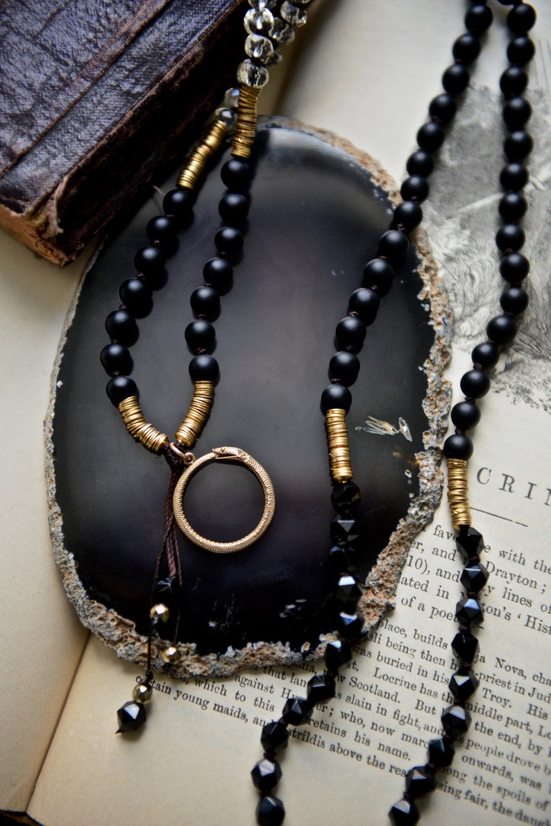 May include: A black and gold beaded necklace with a circular pendant. The necklace is displayed on a grey agate slice and a book with text.