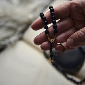 May include: A black beaded necklace with a gold ring pendant. The necklace is held in a hand.