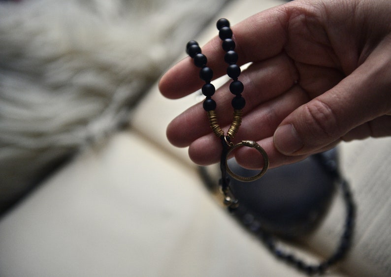 May include: A black beaded necklace with a gold ring pendant. The necklace is held in a hand.