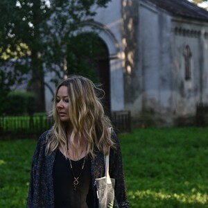 May include: A woman with long blonde hair is standing in front of an old stone building. She is wearing a black top, a long-sleeved patterned jacket, and a tote bag with a fern design. She is also wearing a necklace with a pendant.