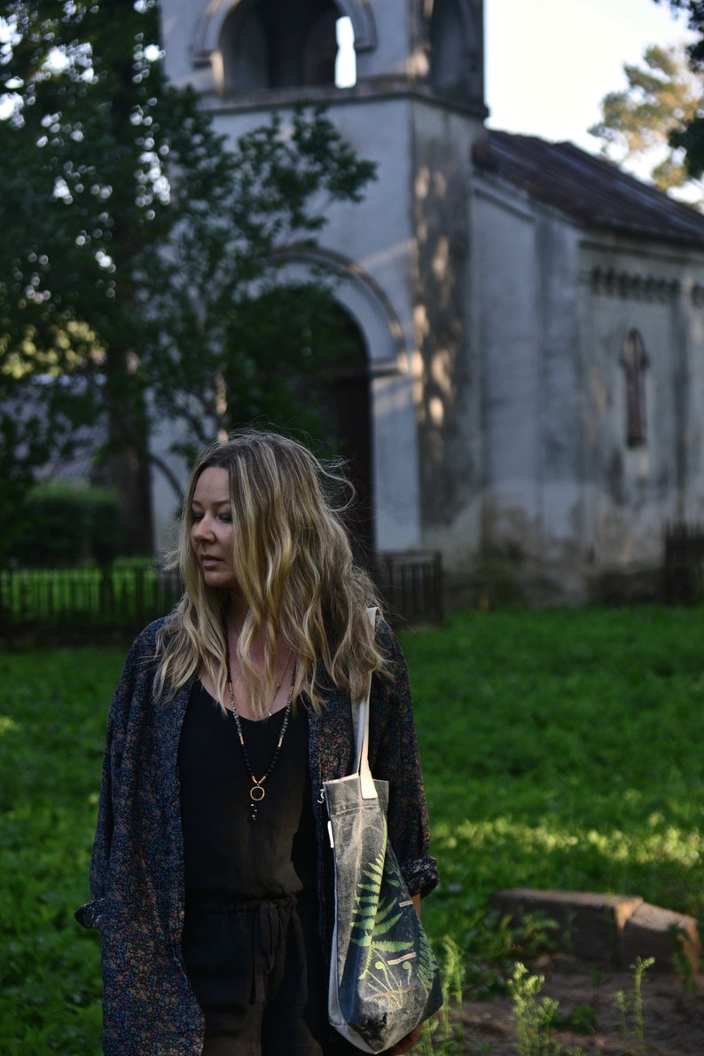 May include: A woman with long blonde hair is standing in front of an old stone building. She is wearing a black top, a long-sleeved patterned jacket, and a tote bag with a fern design. She is also wearing a necklace with a pendant.