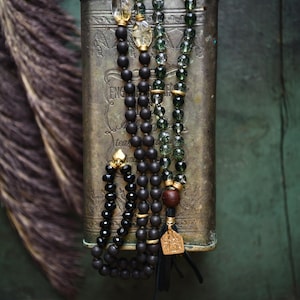 May include: A multi-strand beaded necklace featuring matte black beads, translucent green beads, and clear yellow oval beads. The necklace includes gold-toned metal spacers, a dark brown wooden bead, and a gold-toned pendant with a tassel.