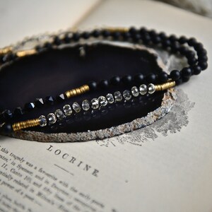 May include: A black and gold beaded necklace with clear crystal beads. The necklace is displayed on a black agate slice.