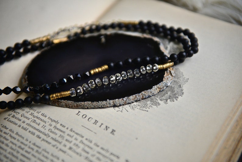 May include: A black and gold beaded necklace with clear crystal beads. The necklace is displayed on a black agate slice.