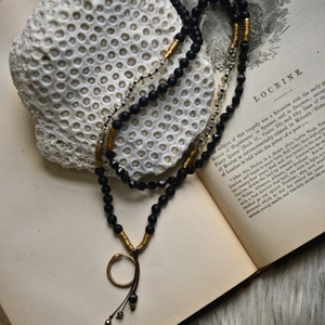 May include: A black and gold beaded necklace with a snake charm pendant. The necklace is laying on an open book with the text "Locrine" visible. The book is on a white furry surface.