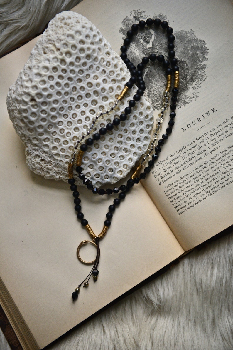 May include: A black and gold beaded necklace with a snake charm pendant. The necklace is laying on an open book with the text "Locrine" visible. The book is on a white furry surface.