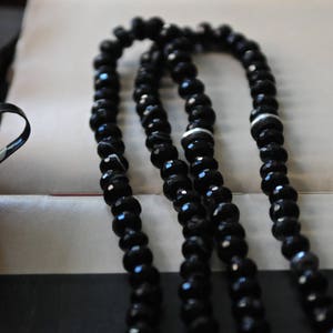 K&ouml;nnte beinhalten: Eine schwarze Perlenkette mit wei&szlig;en Akzenten. Die Kette besteht aus mehreren Perlenstr&auml;ngen und liegt auf einer wei&szlig;en Oberfl&auml;che.