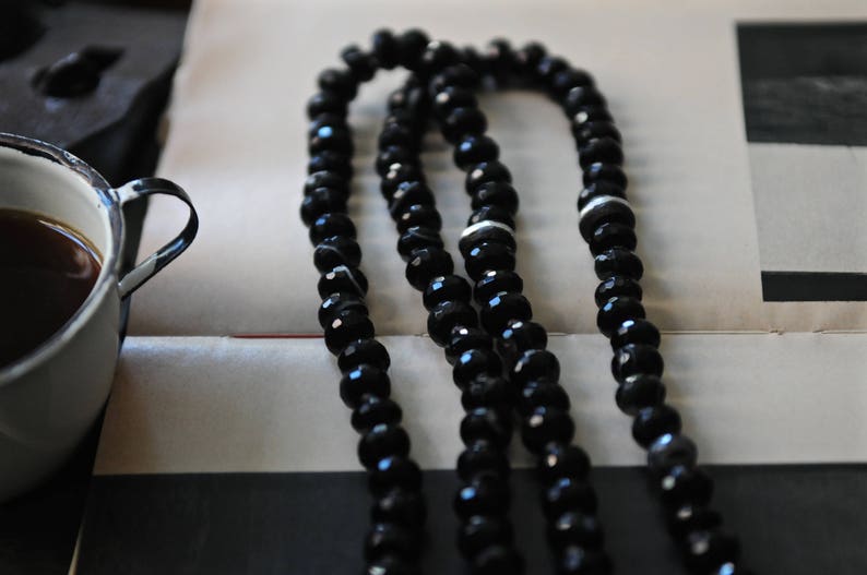 K&ouml;nnte beinhalten: Eine schwarze Perlenkette mit wei&szlig;en Akzenten. Die Kette besteht aus mehreren Perlenstr&auml;ngen und liegt auf einer wei&szlig;en Oberfl&auml;che.