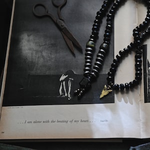 K&ouml;nnte beinhalten: Eine schwarze und goldene Perlenkette mit einem kleinen Pfeil-Anh&auml;nger. Die Kette liegt auf einem Buch mit einem Schwarzwei&szlig;foto einer Person, die auf einem Feld steht. Der Text "... Ich bin allein mit dem Schlagen meines Herzens...  -  Lou Chio" ist auf der Seite gedruckt.