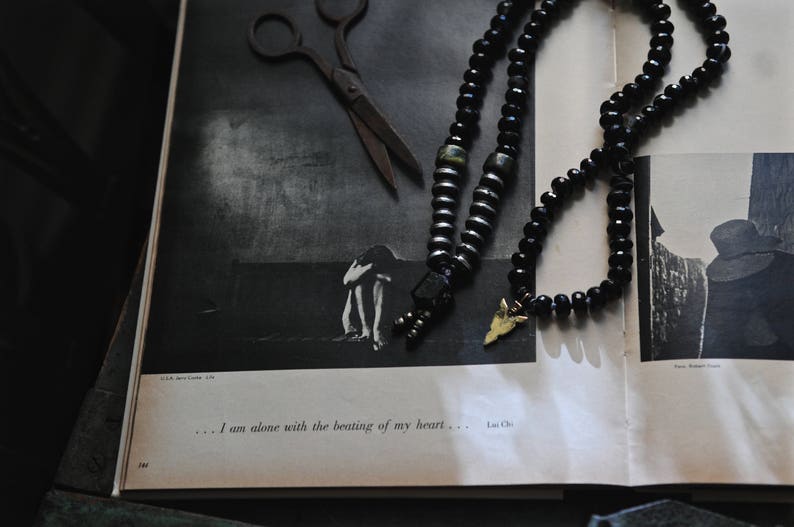K&ouml;nnte beinhalten: Eine schwarze und goldene Perlenkette mit einem kleinen Pfeil-Anh&auml;nger. Die Kette liegt auf einem Buch mit einem Schwarzwei&szlig;foto einer Person, die auf einem Feld steht. Der Text "... Ich bin allein mit dem Schlagen meines Herzens...  -  Lou Chio" ist auf der Seite gedruckt.