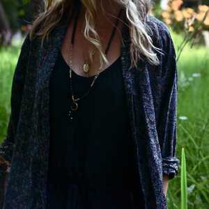 May include: A woman wearing a black tank top with a long-sleeved floral patterned shirt over it. She is wearing a layered necklace with a gold pendant.
