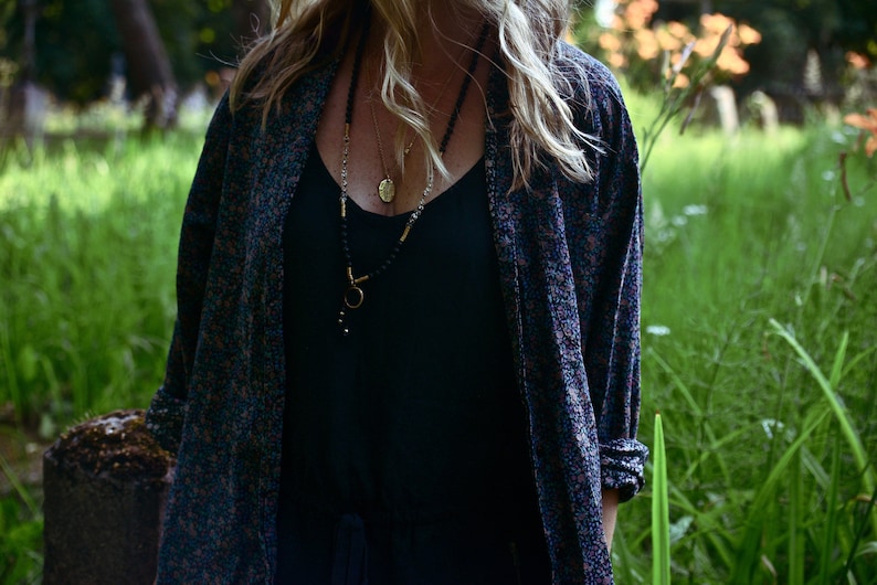 May include: A woman wearing a black tank top with a long-sleeved floral patterned shirt over it. She is wearing a layered necklace with a gold pendant.