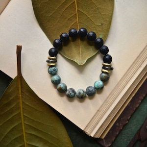 Puede incluir: Una pulsera de cuentas con cuentas azul oscuro y turquesa, acentuada con espaciadores dorados. La pulsera se muestra sobre un libro abierto, con una hoja grande y seca en el fondo. Las cuentas tienen aproximadamente 1,3 cm de diámetro.