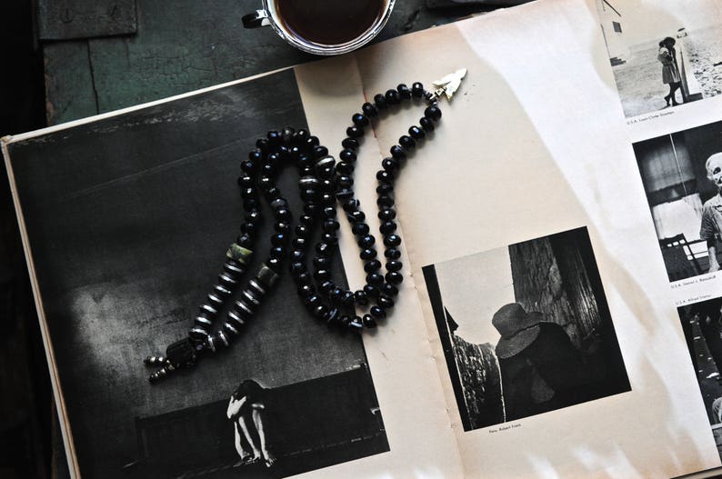 K&ouml;nnte beinhalten: Eine schwarze Perlenkette mit einem silbernen Pfeil-Anh&auml;nger. Die Kette liegt auf einem Buch mit Schwarzwei&szlig;fotos.