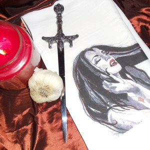 May include: A white kitchen towel featuring a black and white illustration of a vampire woman with long flowing hair. A silver dagger and a clove of garlic are placed on the towel.