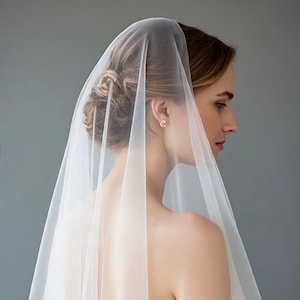 May include: A long, sheer white bridal veil made of fine tulle fabric. The veil is draped over the head and flows down the back, featuring a smooth, unadorned edge. It is worn over a white lace dress with a low back and a row of small fabric buttons.