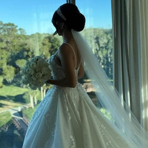 Könnte beinhalten: Eine Braut in einem weißen Brautkleid mit langem Schleier und einem Strauß weißer Rosen steht an einem Fenster und blickt auf eine grüne Landschaft.