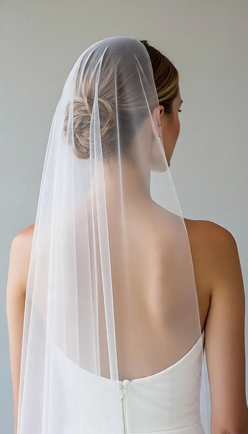 May include: A sheer white bridal veil made of fine tulle fabric, draped elegantly over the back of the head and shoulders. The veil features a smooth, lightweight texture that falls vertically, creating a soft and translucent appearance.