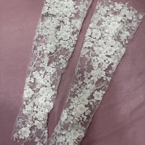 May include: Two white, floral lace sleeves. The sleeves are made of a sheer fabric and are covered in detailed, three-dimensional flower and leaf designs. The sleeves are displayed on a pink background.