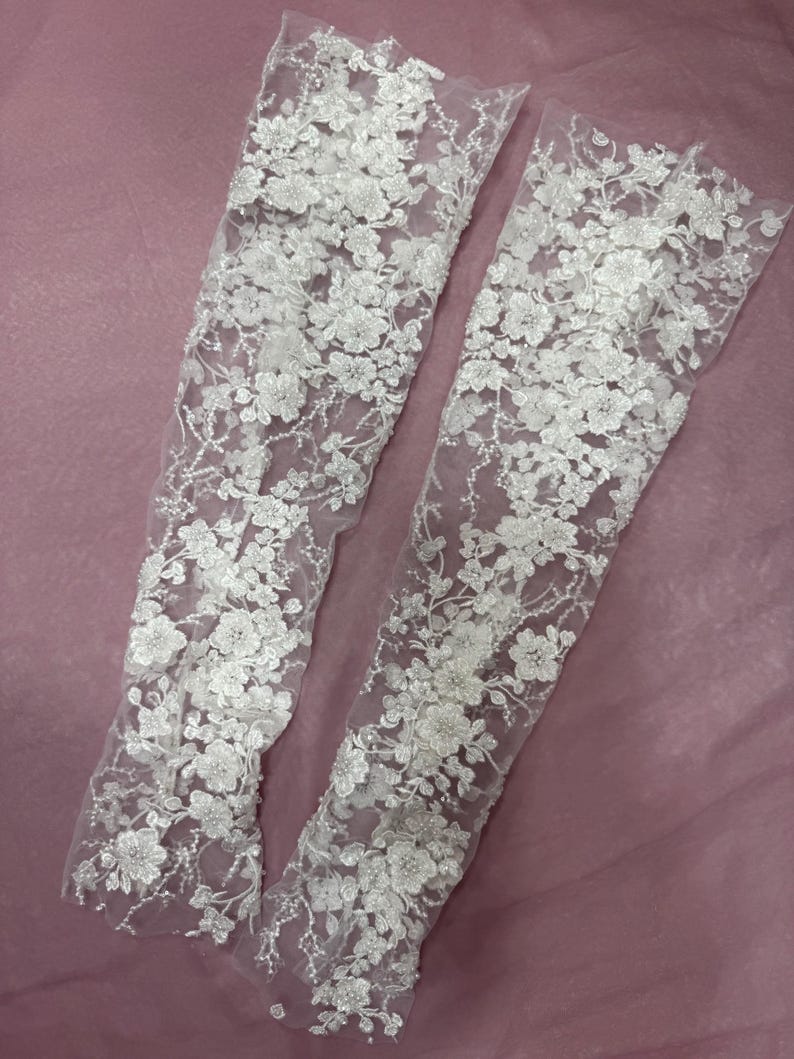 May include: Two white, floral lace sleeves. The sleeves are made of a sheer fabric and are covered in detailed, three-dimensional flower and leaf designs. The sleeves are displayed on a pink background.