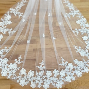 May include: A white bridal veil with a delicate floral lace trim. The lace features 3D flowers and leaves.