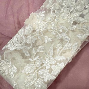 May include: Close-up of white floral lace fabric with intricate embroidered details. The lace features a delicate pattern of flowers and leaves, creating a textured and elegant design. The fabric is likely used for bridal wear or other special occasion garments.
