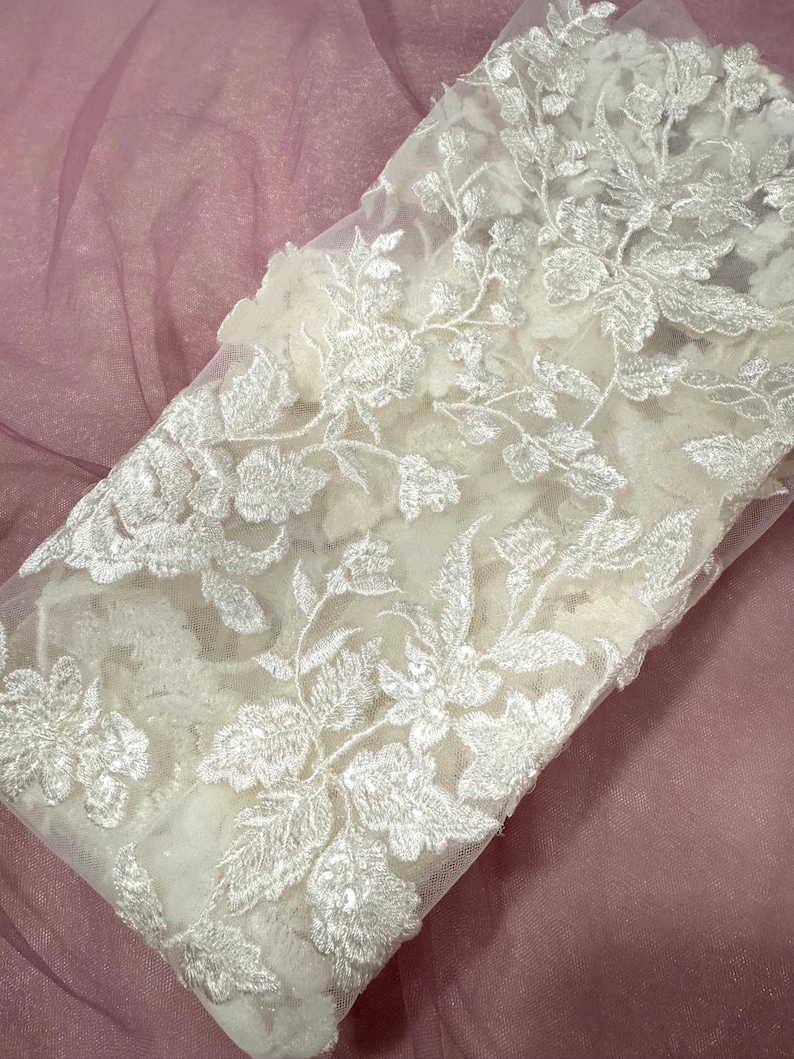 May include: Close-up of white floral lace fabric with intricate embroidered details. The lace features a delicate pattern of flowers and leaves, creating a textured and elegant design. The fabric is likely used for bridal wear or other special occasion garments.