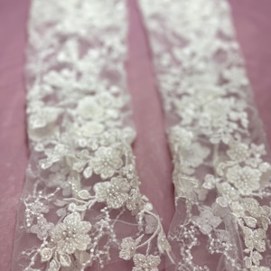 May include: Two white lace arm cuffs with floral and beaded embellishments. The delicate lace features intricate flower patterns and small, sparkling beads. The cuffs are displayed against a soft pink background, highlighting the intricate details.