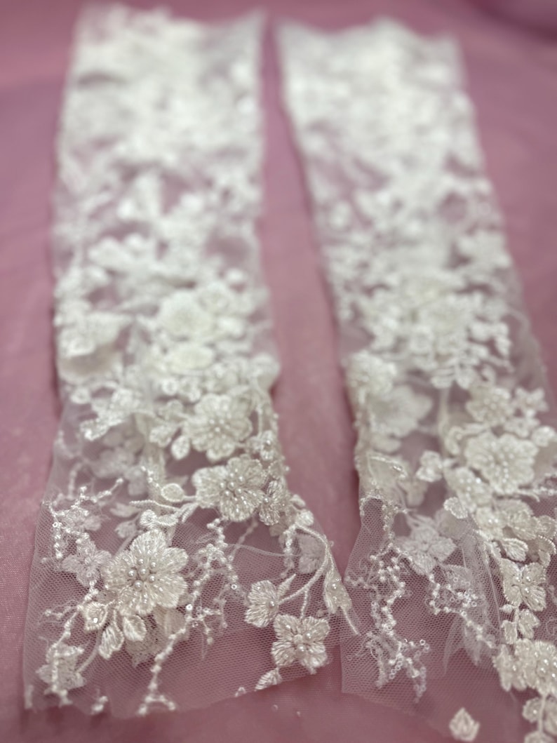 May include: Two white lace arm cuffs with floral and beaded embellishments. The delicate lace features intricate flower patterns and small, sparkling beads. The cuffs are displayed against a soft pink background, highlighting the intricate details.