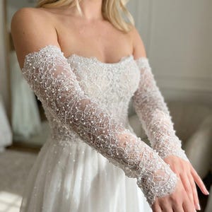 Peut inclure: Une robe de mariée blanche, épaules nues, avec de longues manches en dentelle transparente. Les manches et le corsage sont ornés de perles délicates. La jupe est une conception fluide et superposée, créant un look de mariée romantique et élégant.
