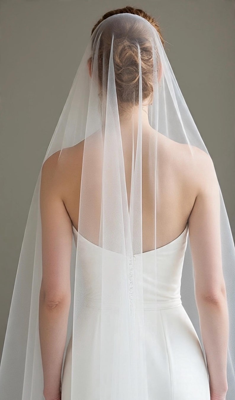 May include: A sheer, floor-length white bridal veil made of fine tulle fabric. The veil is attached to the hair and drapes down the back, extending past the shoulders and torso, creating a soft, layered effect over a strapless white gown.