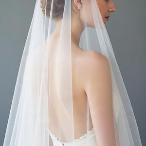 May include: A long, sheer white bridal veil made of fine tulle fabric. The veil is draped over the head and flows down the back, featuring a smooth, unadorned edge. It is worn over a white lace dress with a low back and a row of small fabric buttons.