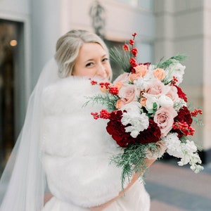 May include: A bride in a white wedding dress with a white fur shawl holds a bouquet of red, pink, and white flowers with greenery and red berries.