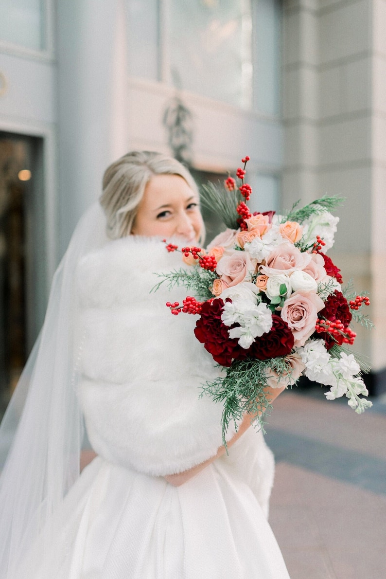 May include: A bride in a white wedding dress with a white fur shawl holds a bouquet of red, pink, and white flowers with greenery and red berries.