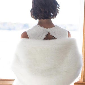 May include: A white faux fur shawl draped over a white dress. The dress has a lace overlay and a keyhole back. The person in the image has short, dark brown hair. The background is a window with a view of a body of water.