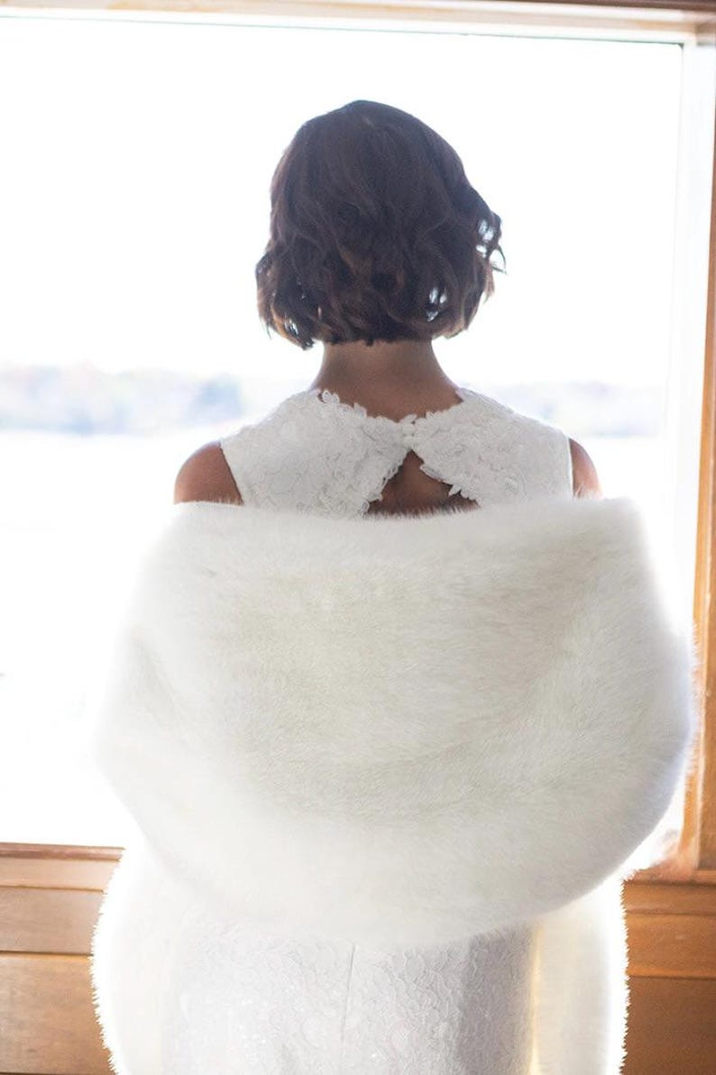May include: A white faux fur shawl draped over a white dress. The dress has a lace overlay and a keyhole back. The person in the image has short, dark brown hair. The background is a window with a view of a body of water.