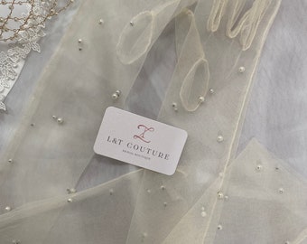 May include: A pair of ivory tulle gloves with pearl embellishments. The gloves are folded and arranged on a white surface. A business card with the text "L&T Couture Bridal Boutique" is visible in the image.
