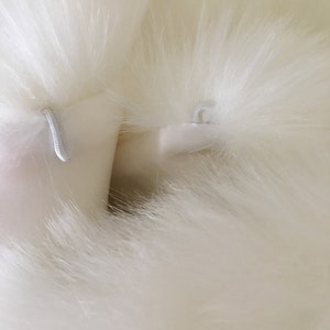 May include: Close-up of a white faux fur fabric with two white loops sewn into the fabric.