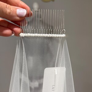 May include: A white tulle veil attached to a silver-toned metal hair comb with thin wire teeth. The veil features a white fabric tag printed with the text L&T COUTURE BRIDAL BOUTIQUE. The comb is held at the top, showcasing the fine mesh fabric.
