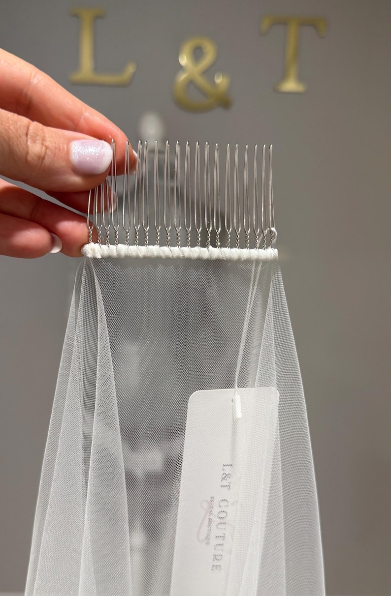 May include: A white tulle veil attached to a silver-toned metal hair comb with thin wire teeth. The veil features a white fabric tag printed with the text L&T COUTURE BRIDAL BOUTIQUE. The comb is held at the top, showcasing the fine mesh fabric.