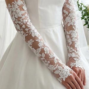 May include: White lace bridal gloves with floral patterns. The gloves are long-sleeved and made of delicate, sheer fabric. The intricate floral design covers the entire length of the gloves, creating an elegant and romantic look. The gloves are paired with a white wedding dress.