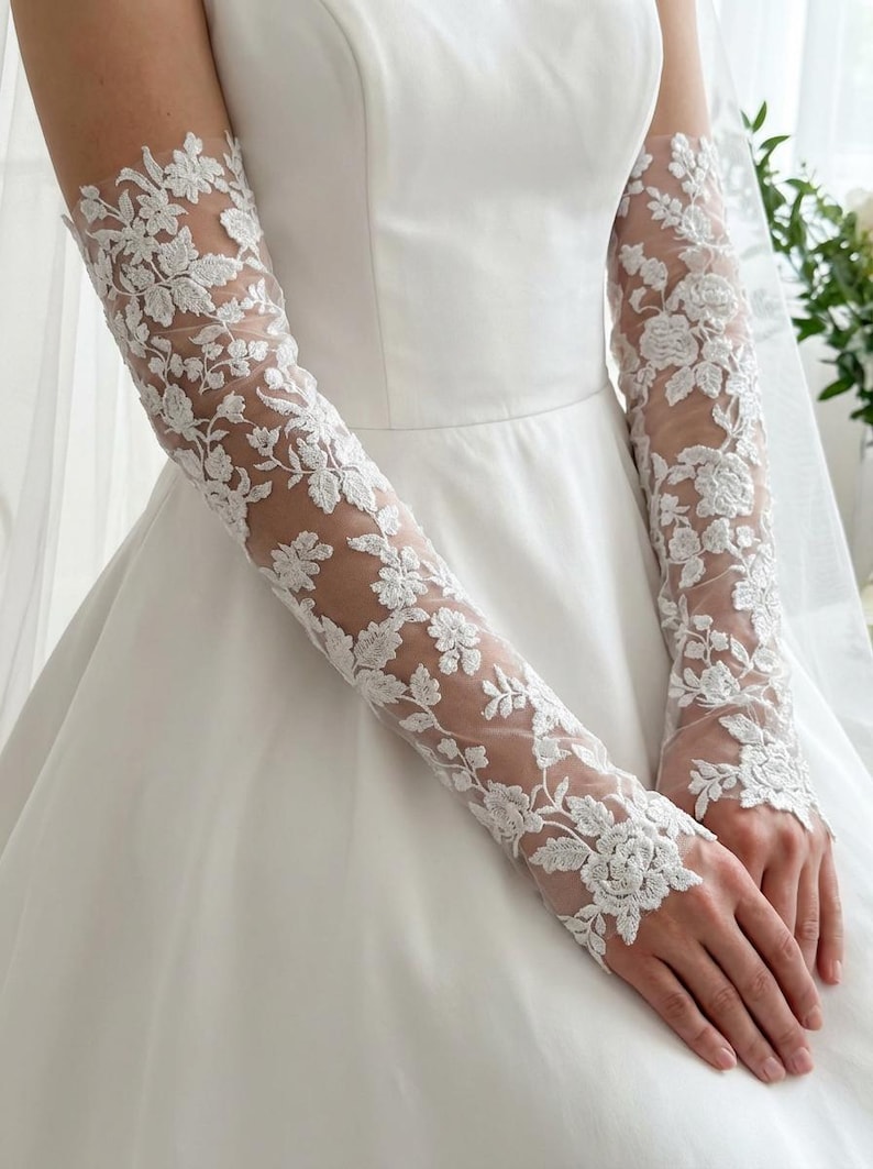 May include: White lace bridal gloves with floral patterns. The gloves are long-sleeved and made of delicate, sheer fabric. The intricate floral design covers the entire length of the gloves, creating an elegant and romantic look. The gloves are paired with a white wedding dress.