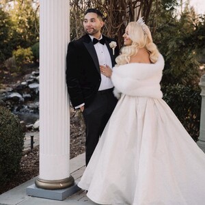May include: A bride and groom pose for a wedding portrait. The bride is wearing a white gown with a fur shawl and a tiara. The groom is wearing a black tuxedo with a velvet jacket. They are standing in front of a white pillar.