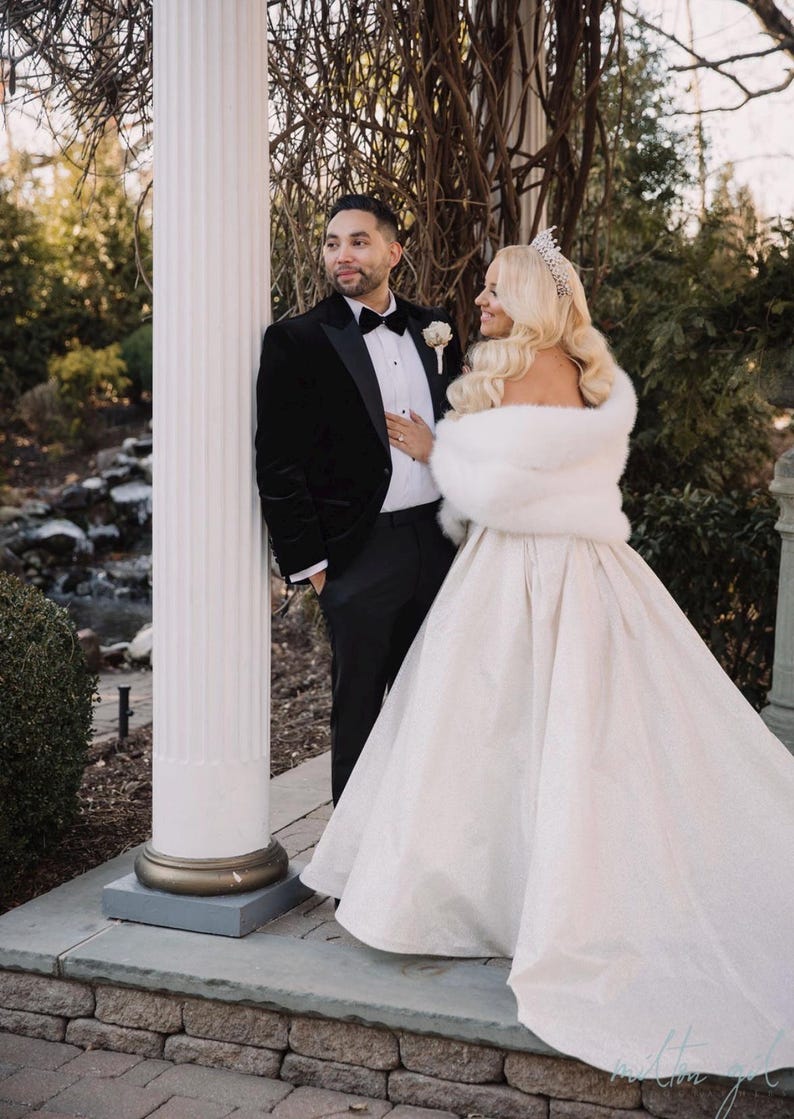May include: A bride and groom pose for a wedding portrait. The bride is wearing a white gown with a fur shawl and a tiara. The groom is wearing a black tuxedo with a velvet jacket. They are standing in front of a white pillar.
