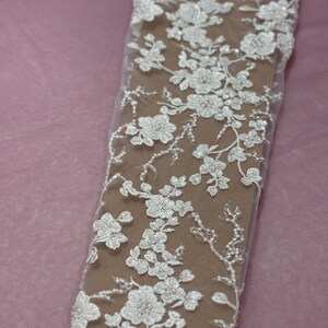 May include: Close-up of white floral lace fabric. The lace features detailed, three-dimensional flower and leaf patterns with beaded accents. The fabric is set against a neutral background, highlighting the delicate design.