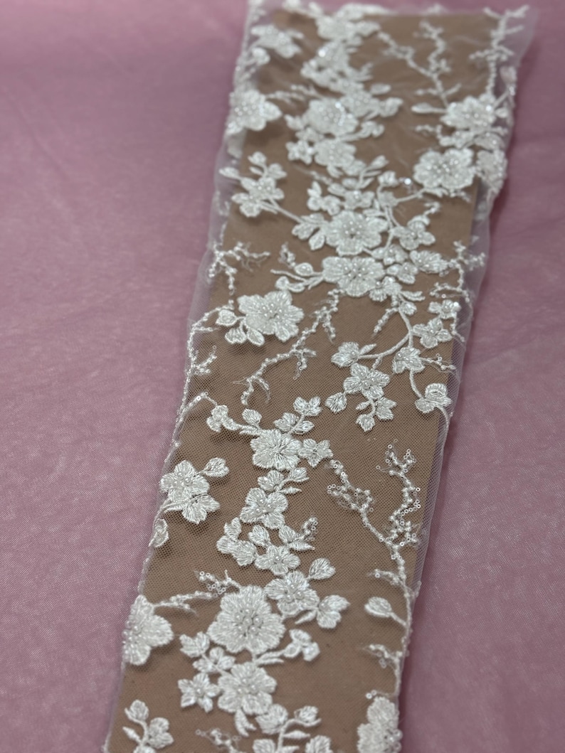 May include: Close-up of white floral lace fabric. The lace features detailed, three-dimensional flower and leaf patterns with beaded accents. The fabric is set against a neutral background, highlighting the delicate design.