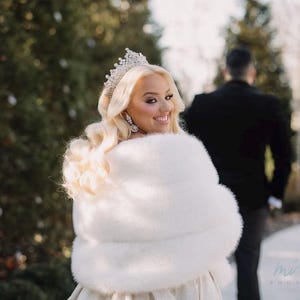 May include: A bride wearing a white fur shawl and a tiara, looking over her shoulder at the groom, who is standing behind her. The bride is smiling and the groom is wearing a black suit.