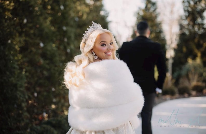 May include: A bride wearing a white fur shawl and a tiara, looking over her shoulder at the groom, who is standing behind her. The bride is smiling and the groom is wearing a black suit.